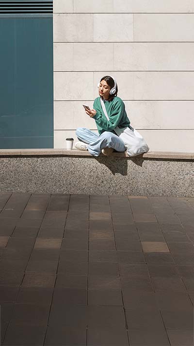 Eine junge Frau sitzt im Schneidersitz auf einer niedrigen Mauer und schaut auf Ihr Smartphone. 