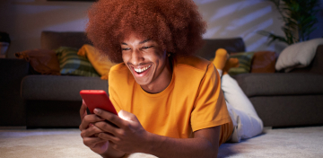 Eine Person mit einem breiten Lächeln liegt auf dem Bauch in einem gemütlichen Wohnzimmer und hält ein rotes Smartphone in den Händen. Im Hintergrund sind ein graues Sofa mit bunten Kissen und eine Zimmerpflanze zu sehen. 