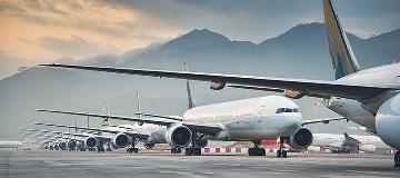 Viele Flugzeuge stehen geparkt in einer Reihe auf einem Rollfeld. Dahinter erheben sich wolkenverhangene Berge.